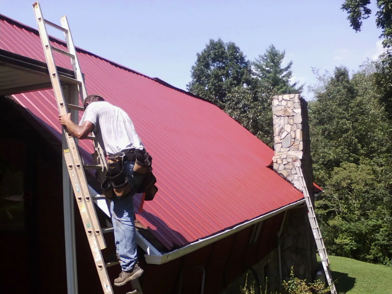 Roofer on a ladder beside a red metal roof and stone chimney — Soffit & Fascia Repair by Roofing Brothers in Central Point